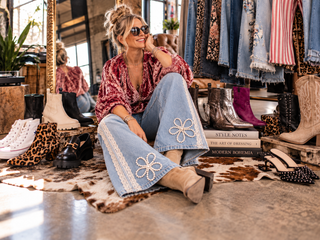 Stylish woman wearing embellished wide-leg jeans and western boots surrounded by statement footwear in a colourful boho-inspired fashion studio.