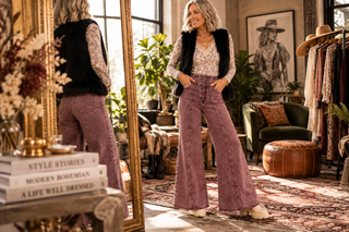 Stylish silver-haired woman adjusting a blouse tucked into high-waisted wide-leg jeans in a glamorous bohemian dressing room.